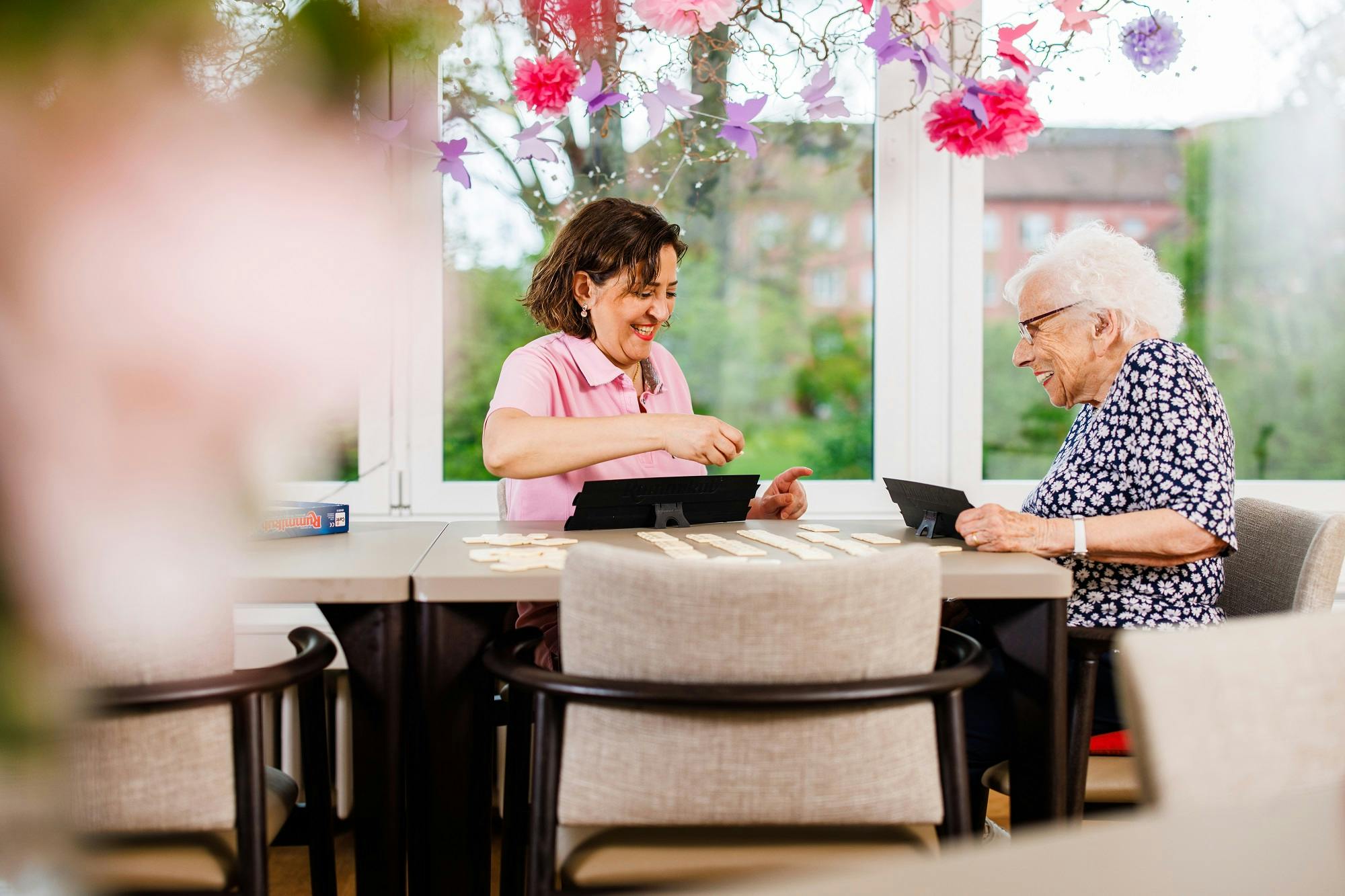Jüngere Frau und ältere Frau lächeln beim Betrachten eines Tablets in einem hellen Raum mit Blumendekoration.